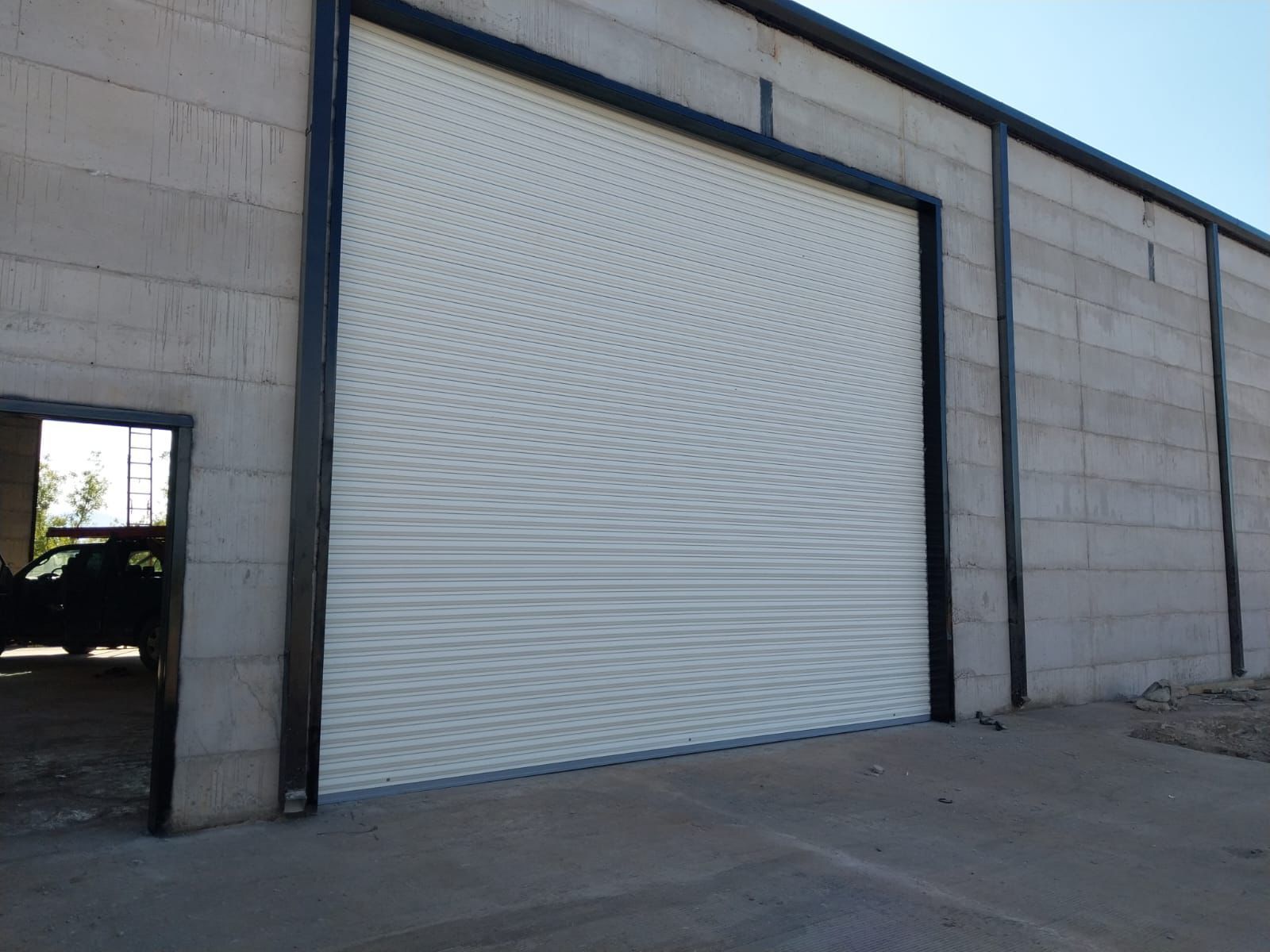Una gran puerta enrollable de color blanquecino en un edificio de hormigón con molduras negras. Una puerta más pequeña se abre a la izquierda.