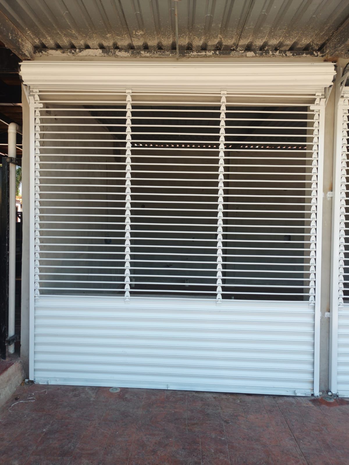 Puerta de seguridad blanca con lamas horizontales y base de metal corrugado. Bajo un toldo de metal corrugado.