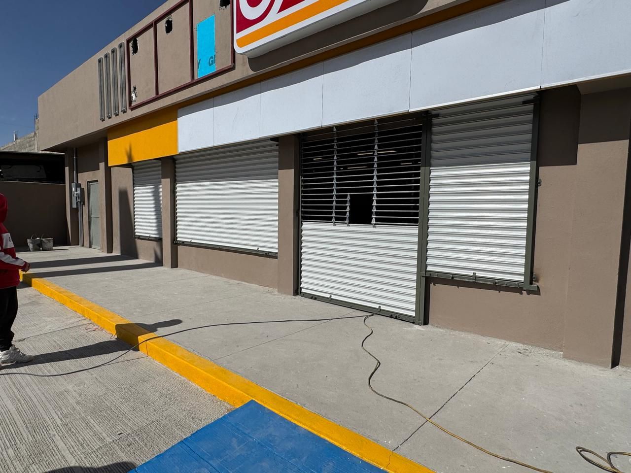 Exterior de un negocio con persianas de seguridad blancas cerradas, edificio amarillo y marrón, y una persona parada cerca de la acera.