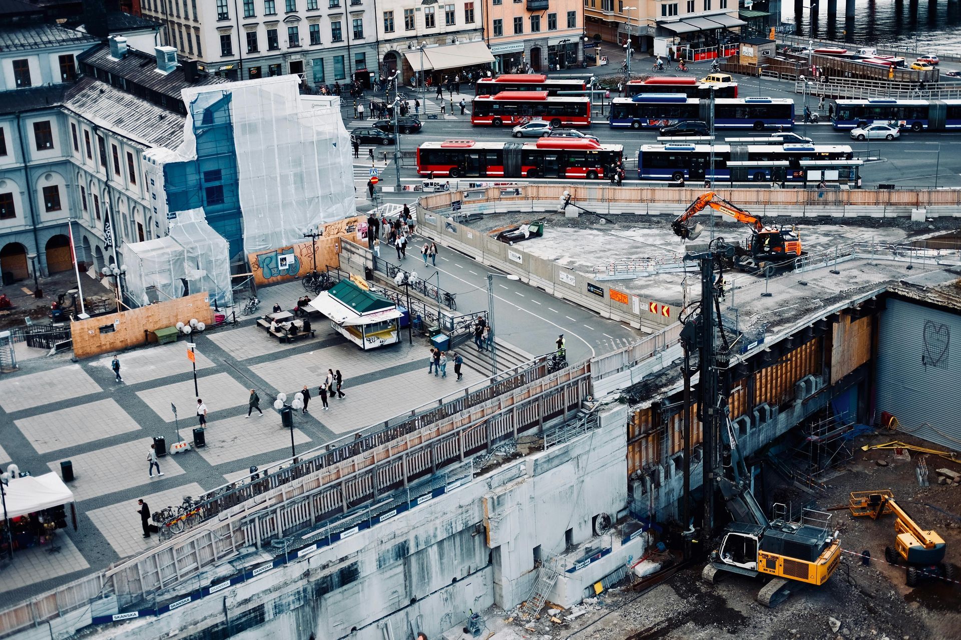 Construction site in a city square, with workers, excavators, buses, and surrounding buildings.