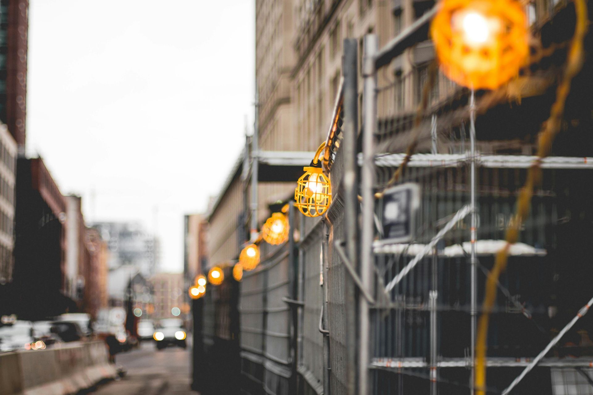 String lights decorate a construction barrier on a city street. Warm yellow globes illuminate the grey metal fencing.