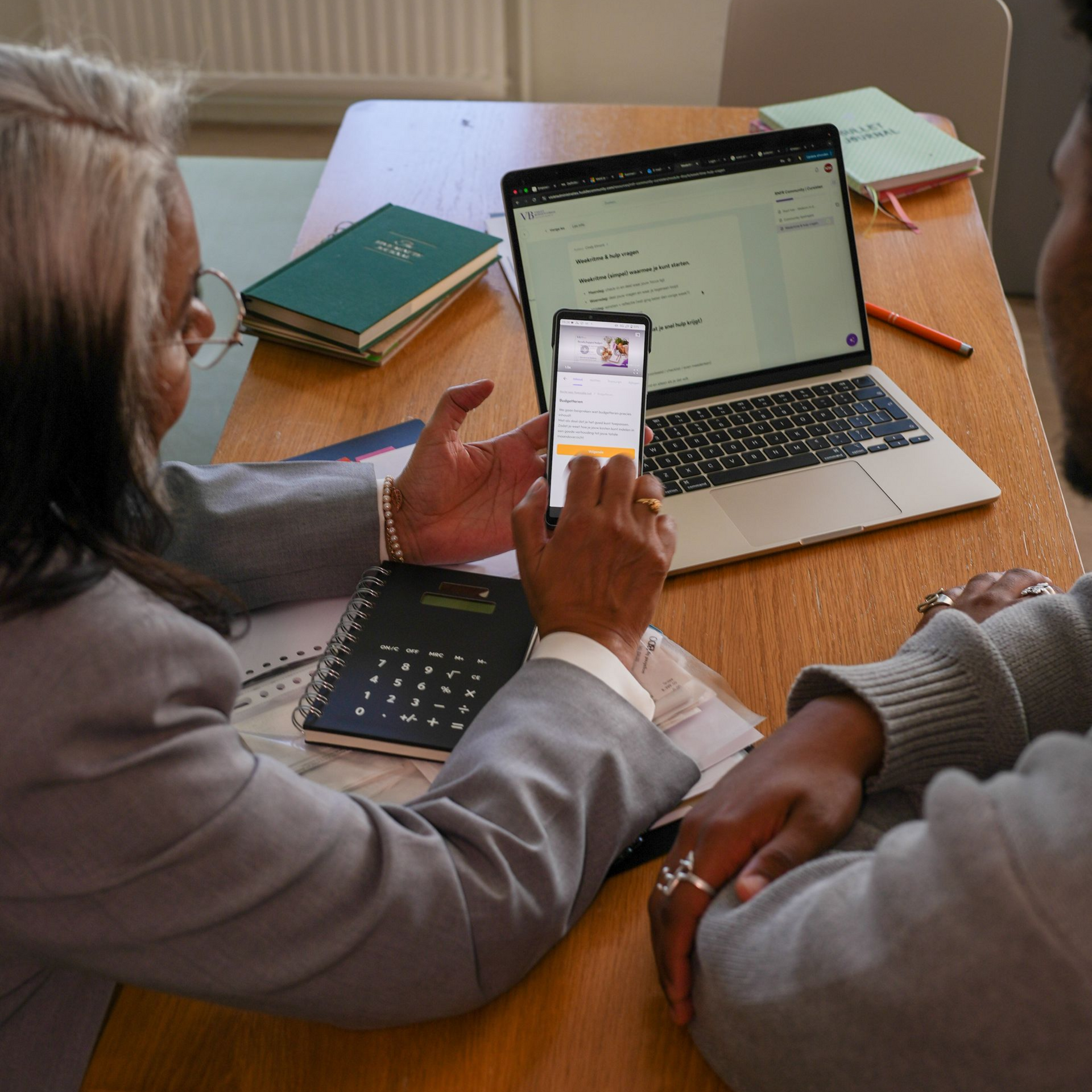 Mensen die een smartphone bekijken naast een opengeklapte laptop op een houten tafel met notitieboekjes en pennen.