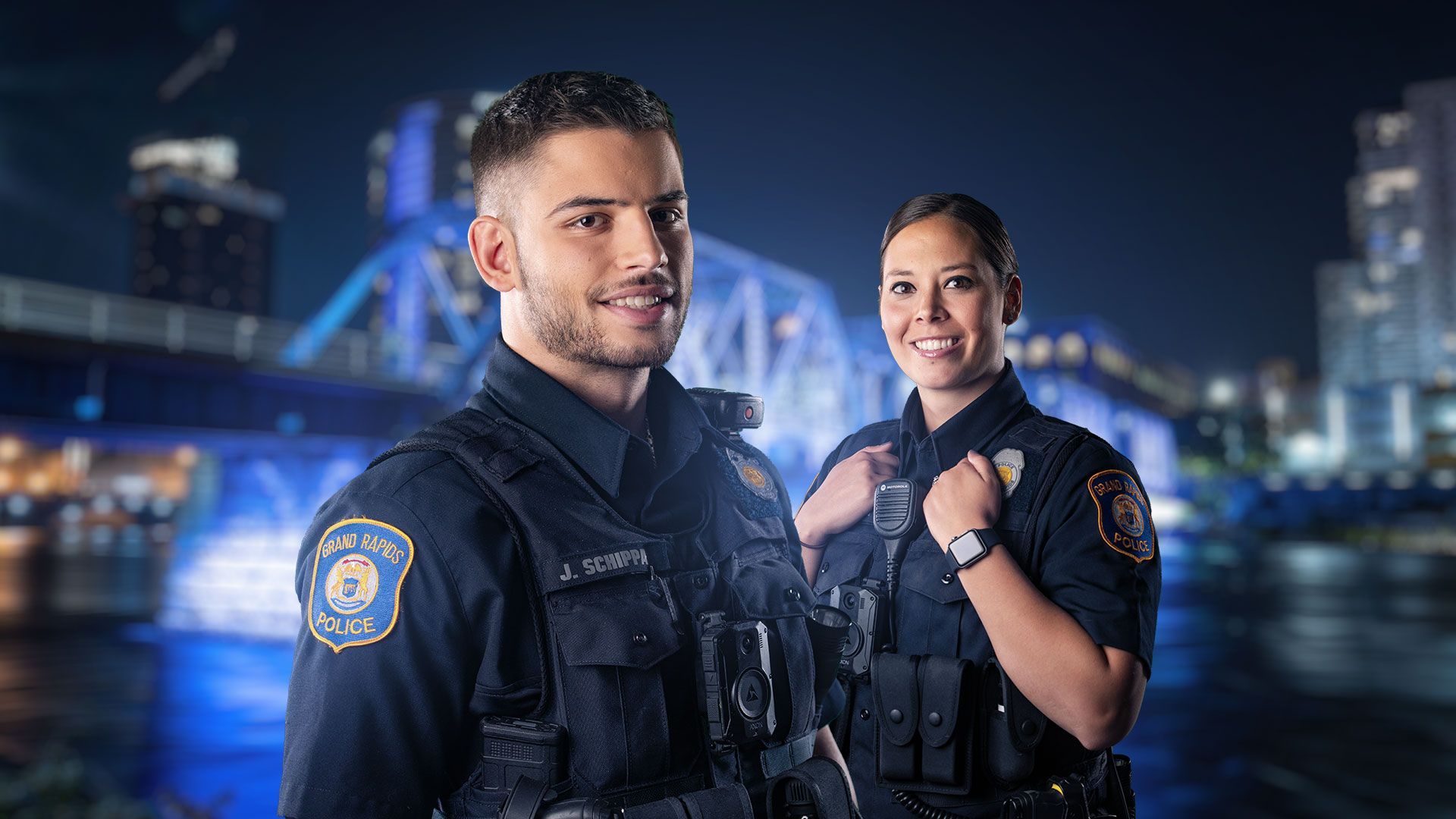 Two uniformed police officers smiling in front of a blue-lit bridge at night.