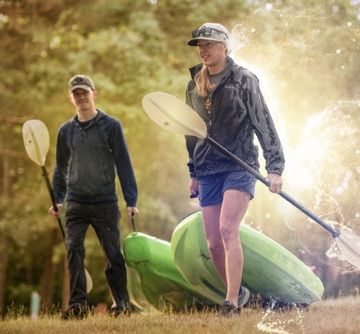 Two people with paddles stand near two green kayaks in a grassy area.
