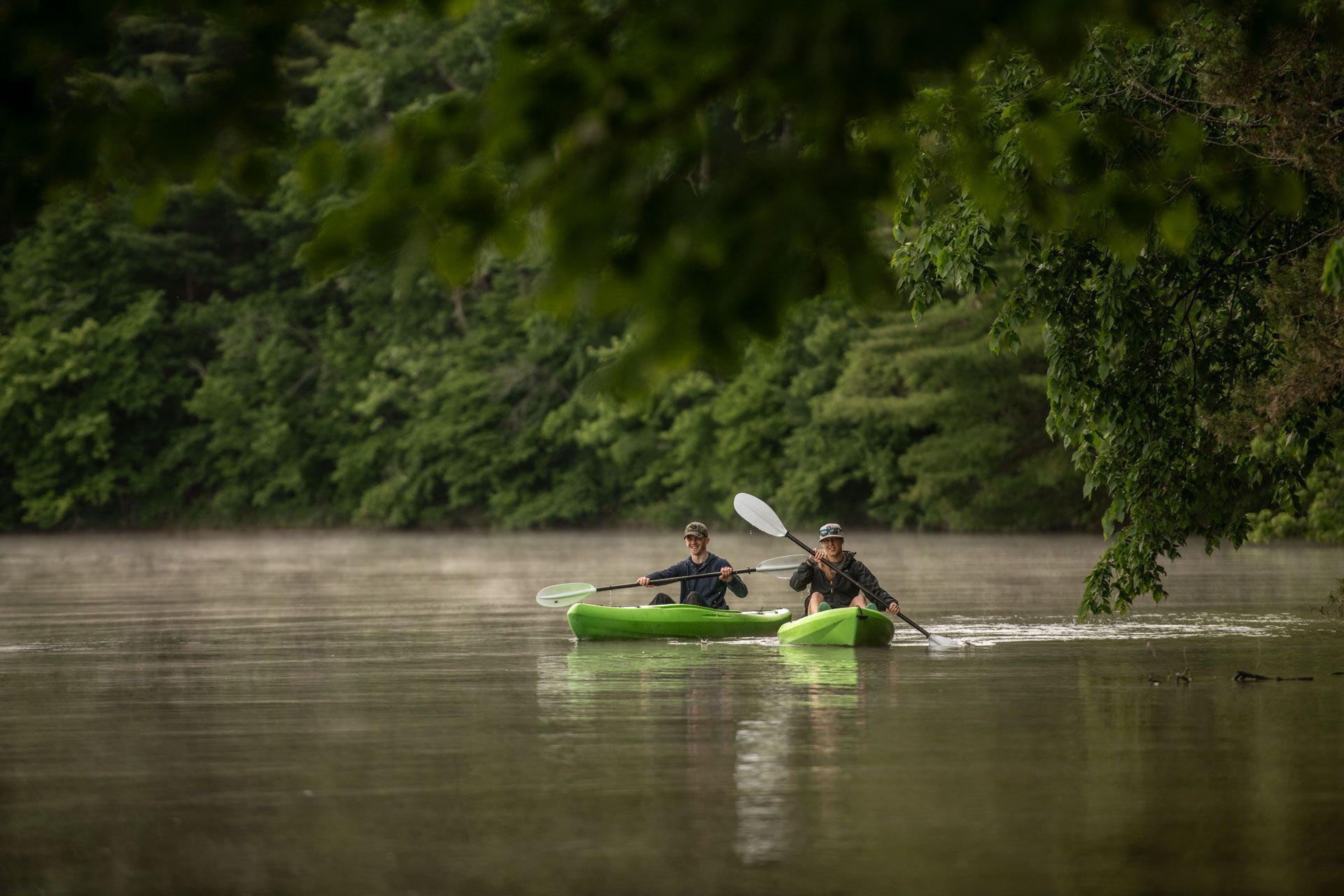 Two people kayaking on a misty river, surrounded by green trees.