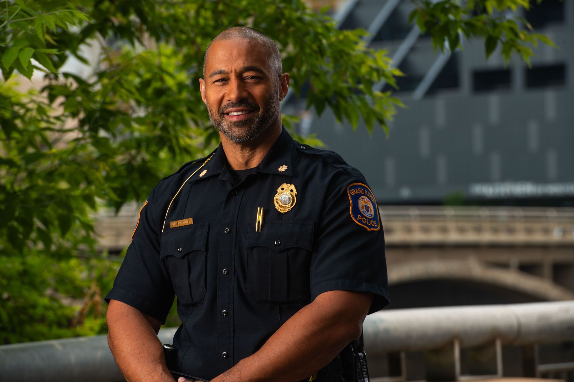 Police officer in dark blue uniform smiling, badge visible, outside near greenery and a building.