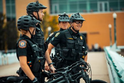 Four police officers in black uniforms and helmets stand near their bicycles on a bridge; one smiles.