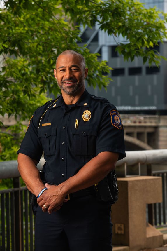 Police officer in uniform smiles against a backdrop of a building and greenery.