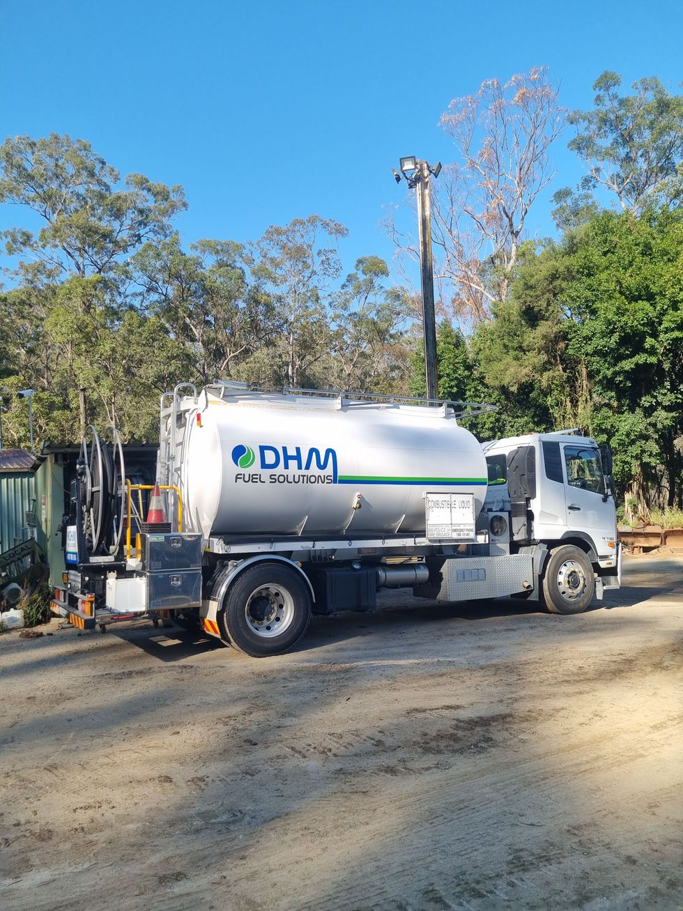 White DHM septic truck parked on a dirt lot, trees in background, blue sky.