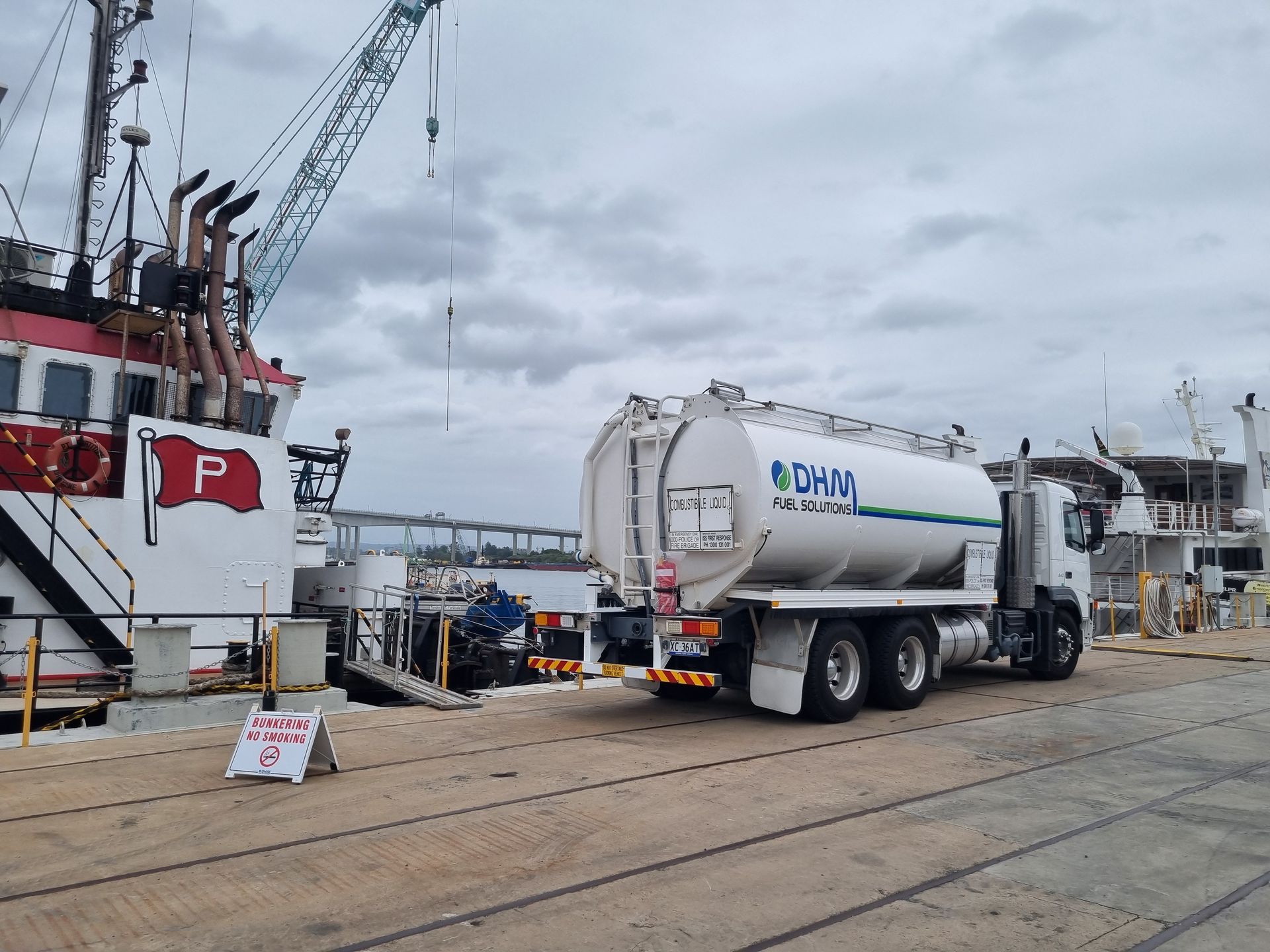 Tanker truck at an industrial dock beside a red-and-white ship under an overcast sky.