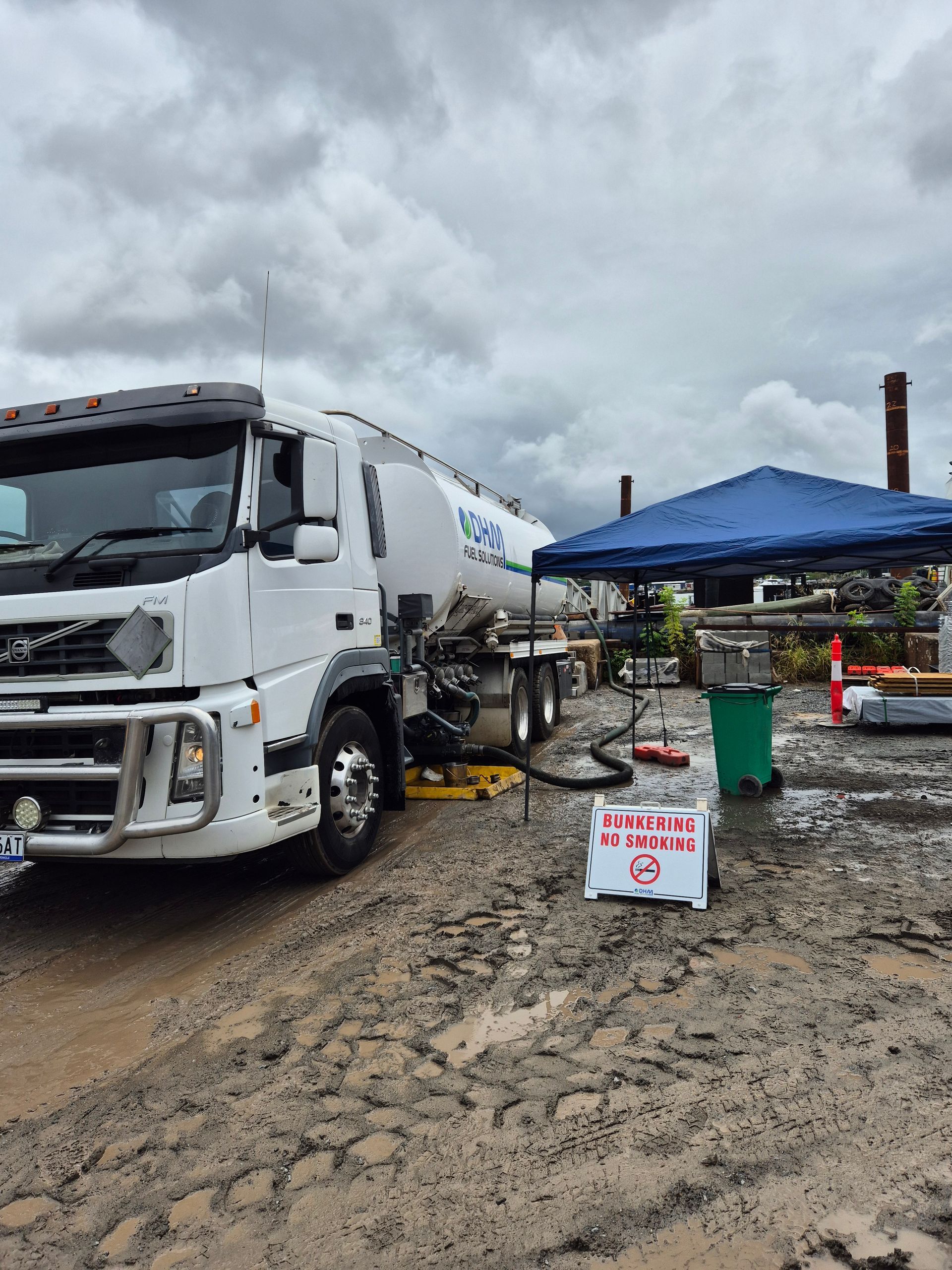 White tanker truck at a muddy worksite beside a blue canopy and a small sign under cloudy skies