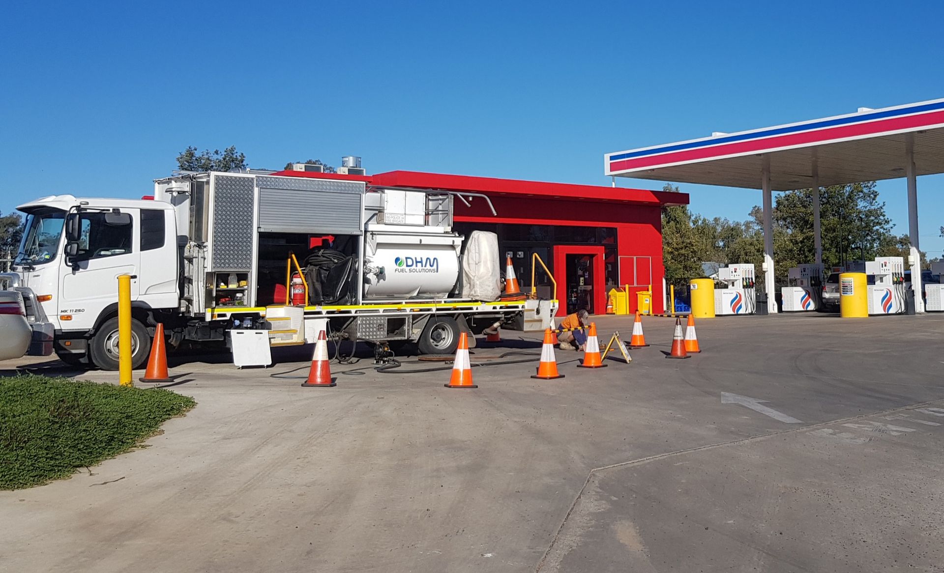 White truck with equipment at a gas station, surrounded by orange traffic cones.