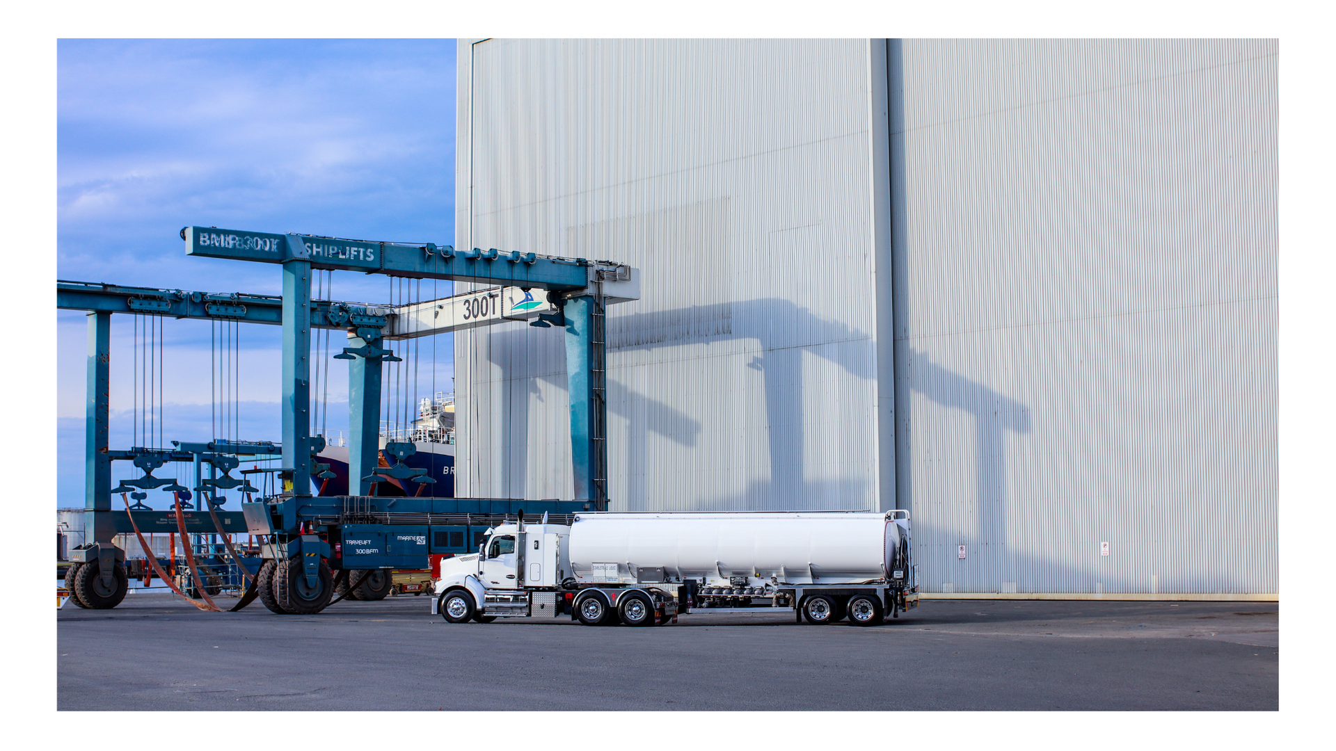 A white tanker truck parked in front of a large, blue industrial straddle carrier next to a tall, corrugated warehouse.