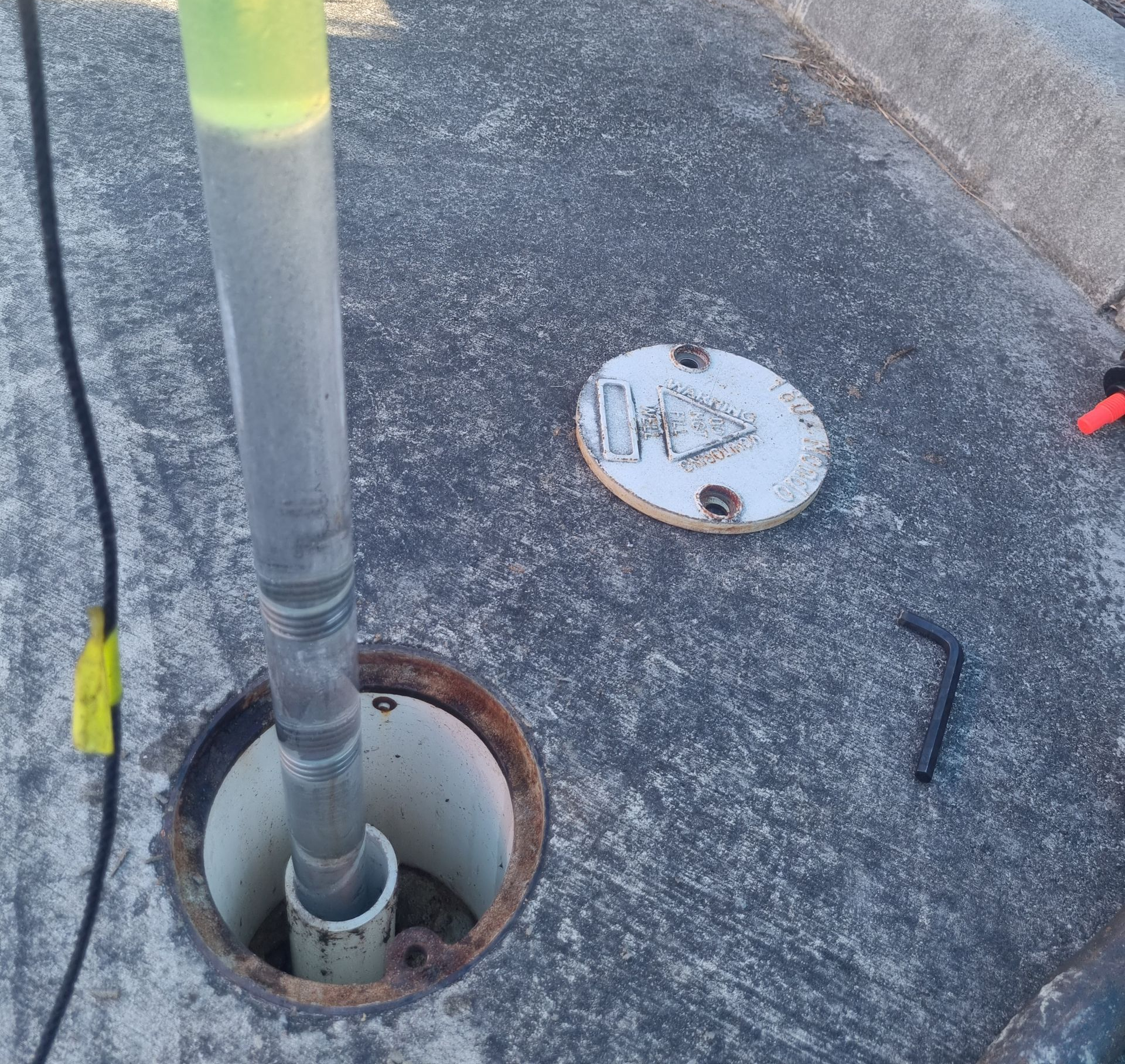 Open utility access hole with a clear vertical pipe and nearby round cover on rough pavement.