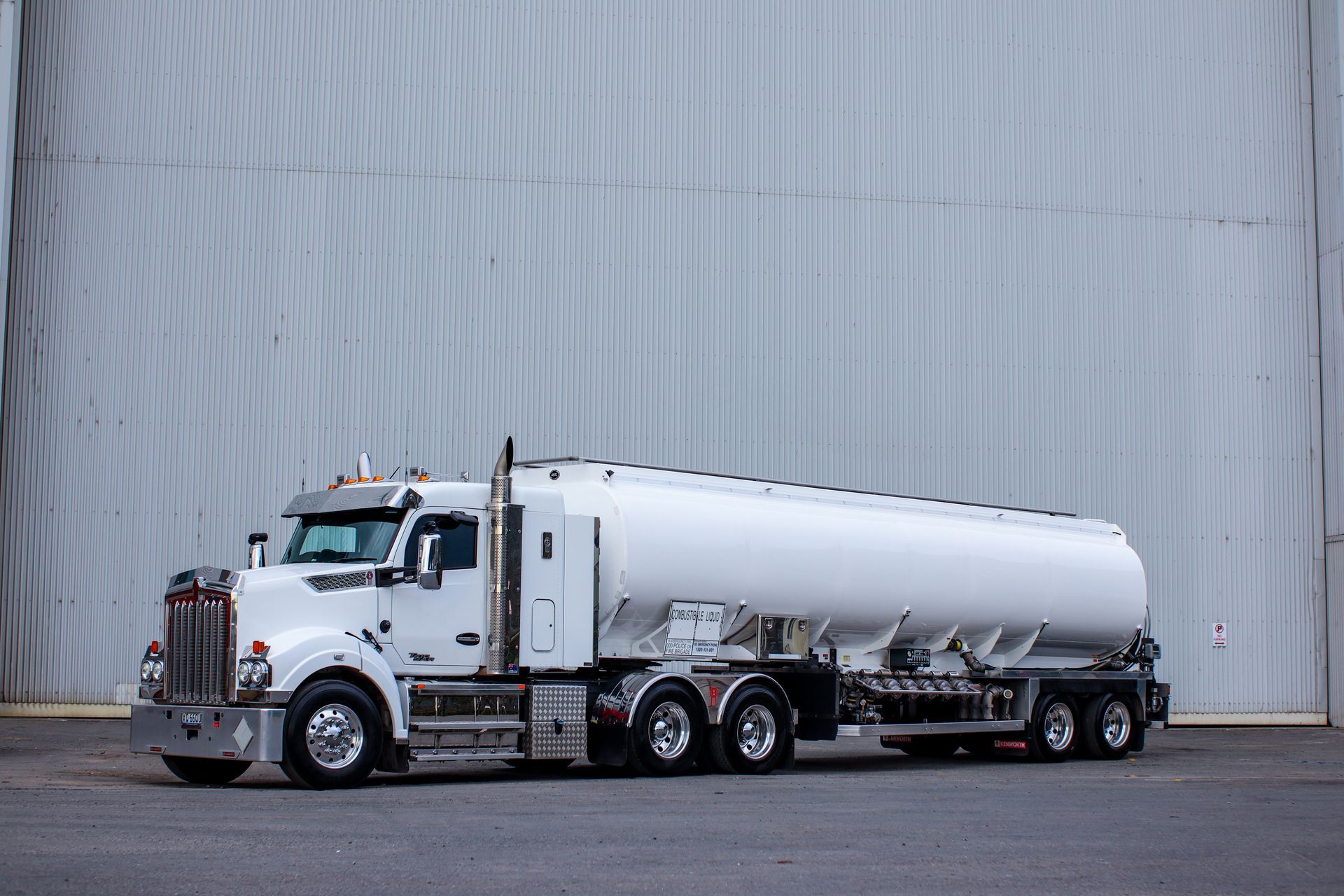 A white semi-truck with a large tanker trailer parked in front of a corrugated metal wall.