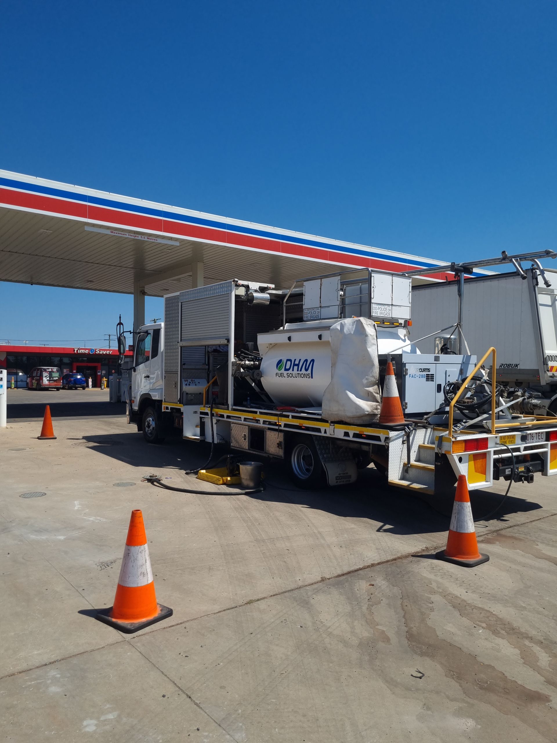 A white truck parked under a gas station awning with cones, equipment, and a blue sky.