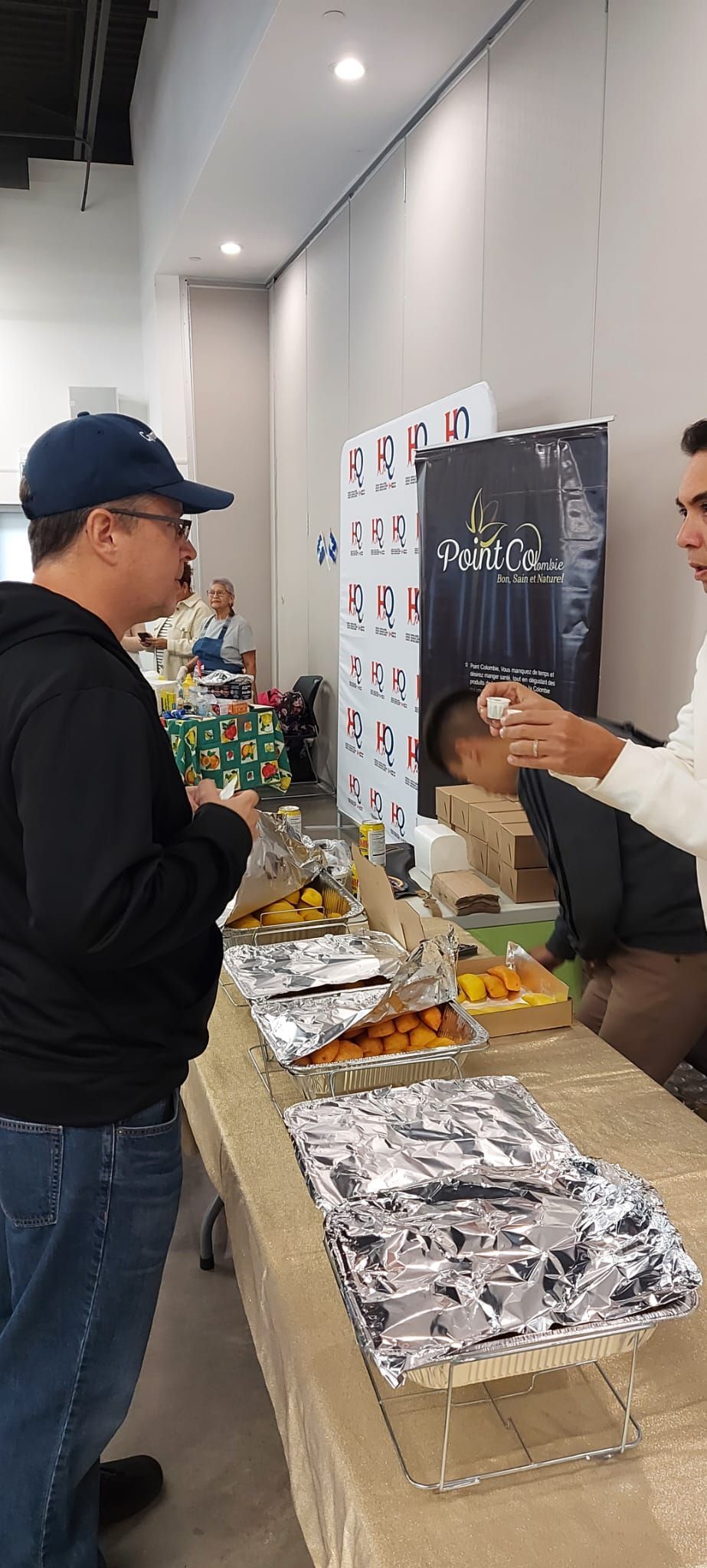 Des personnes attablées à un buffet, l'une coiffée d'une casquette bleue, une autre servant les plats. Nourriture emballée dans des barquettes en aluminium, ambiance d'un événement.