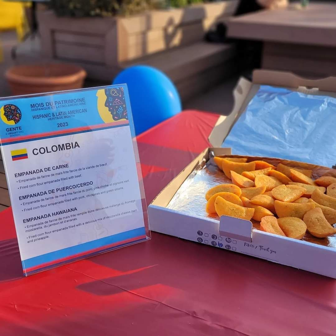 Des empanadas colombiennes et une pancarte sur une table rouge. Un ballon bleu et un mobilier de terrasse sont visibles.