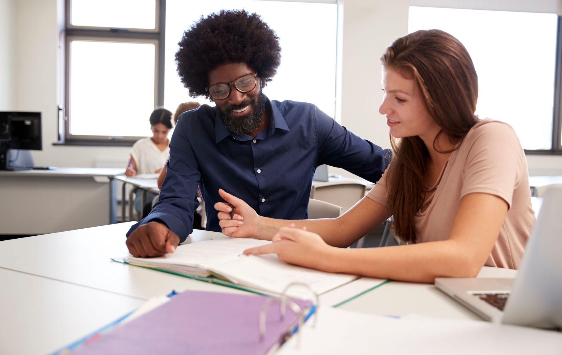 Instructor assisting student with a notebook at a desk in a classroom setting.