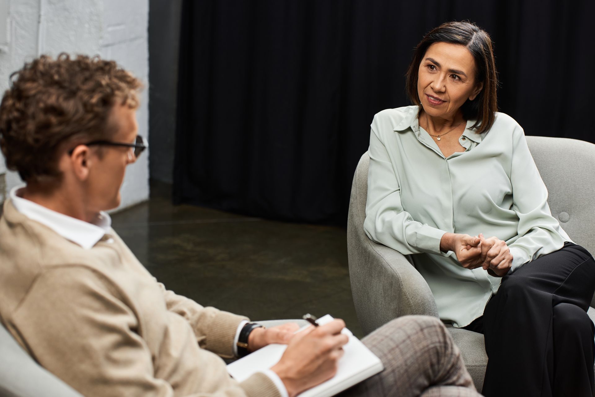 Person in light-green shirt and black pants sits and talks to a person holding a notebook. Both in a room with black curtains.