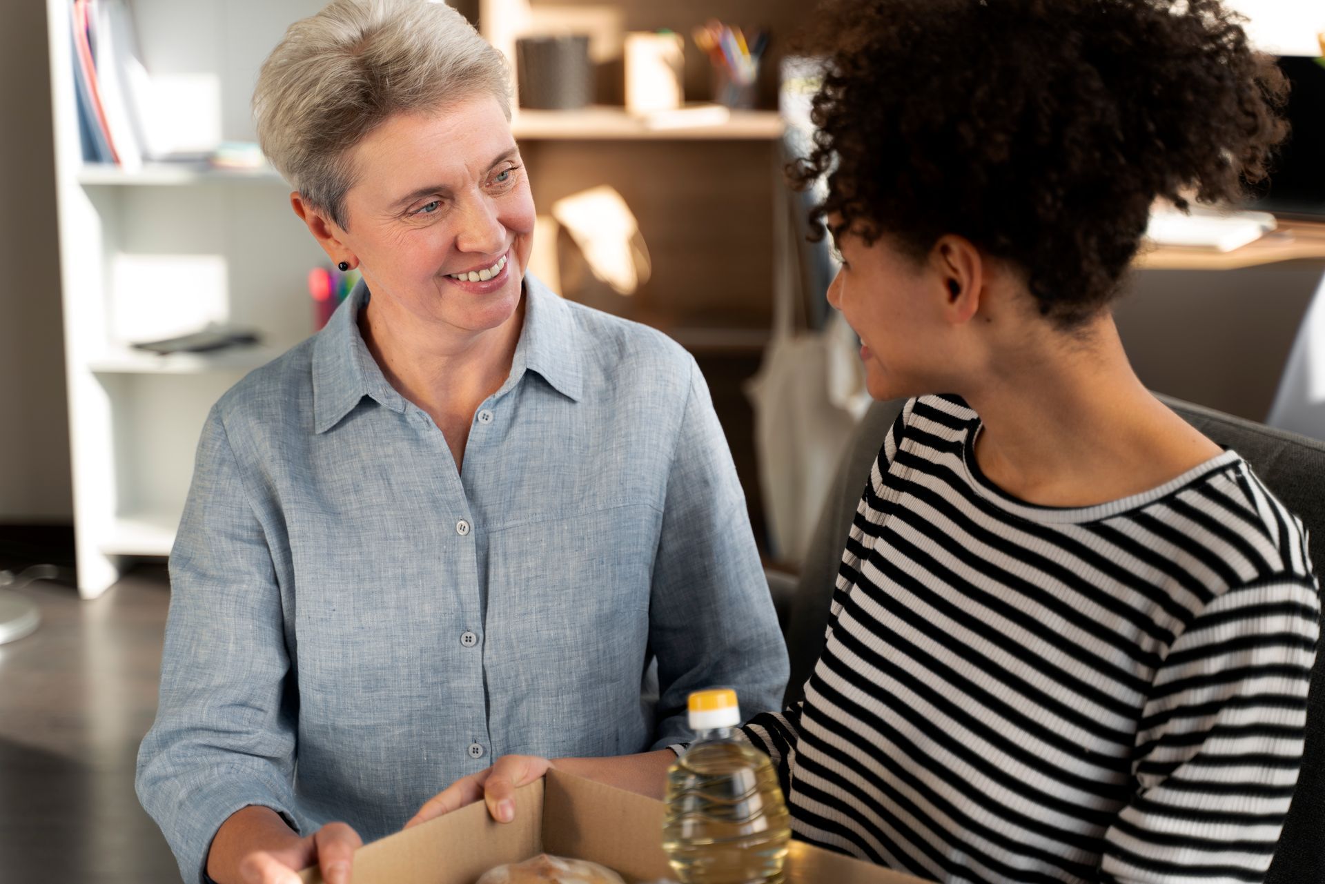 Two people looking at each other, holding a box of food; indoors, sunny.