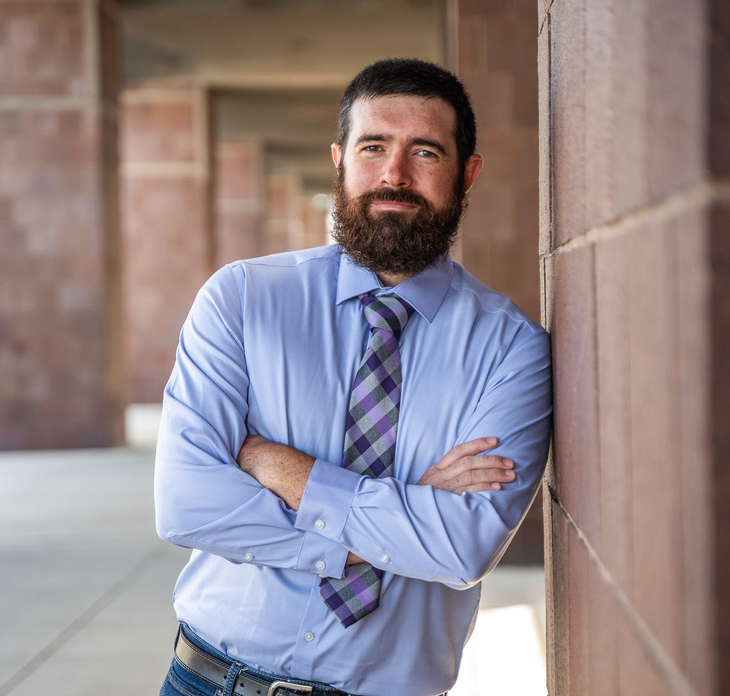Man with beard in blue shirt and tie leaning against a brick wall, arms crossed, outdoors.