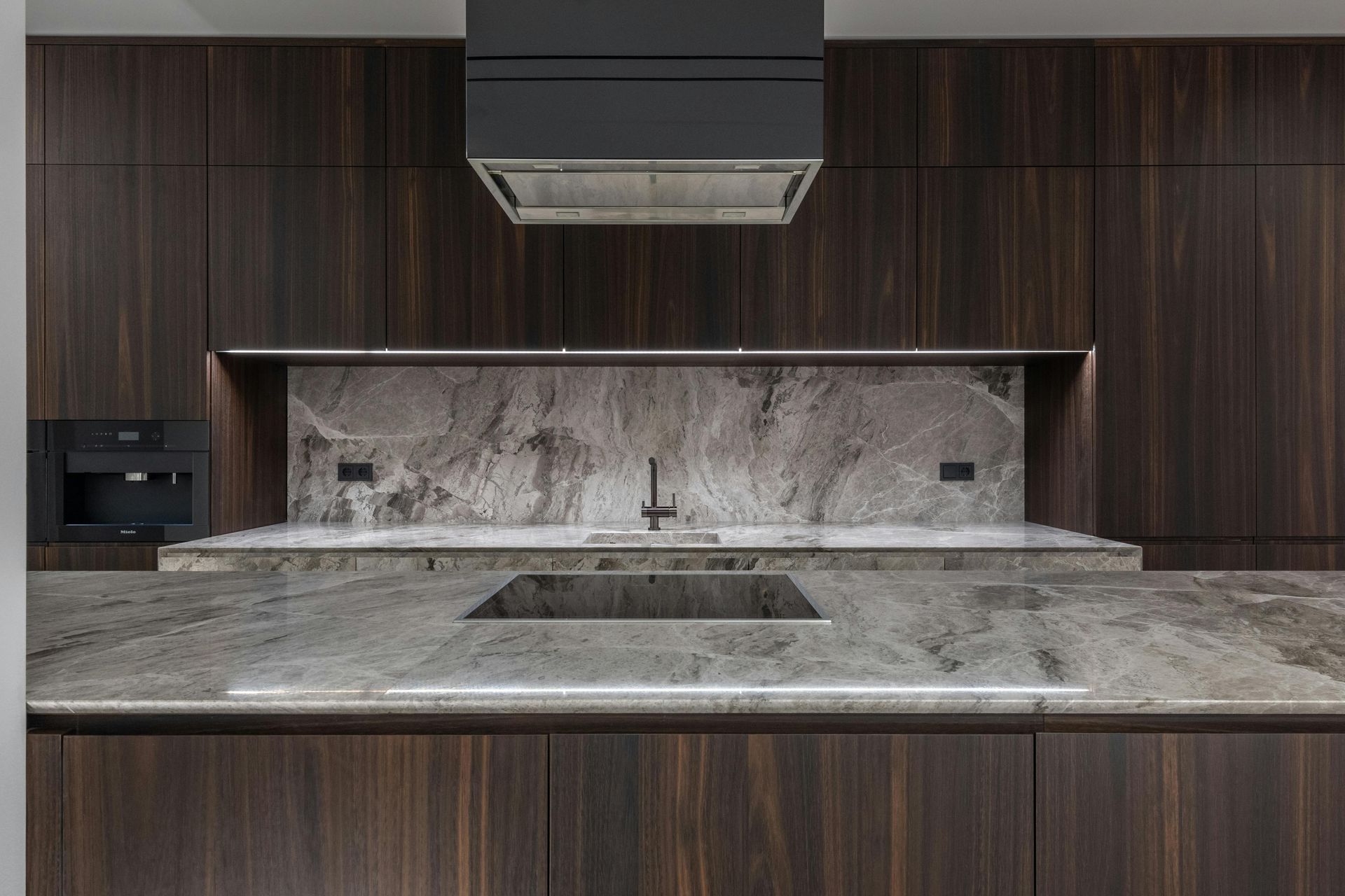 Dark wood kitchen with granite countertops and a built-in sink; black range hood and coffee machine.