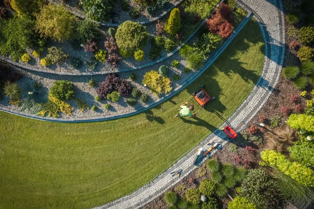 Aerial view of a garden with curved stone pathways, diverse shrubs, and two people mowing the lawn with riding mowers. — Rohan's Maintenance in Robina, QLD