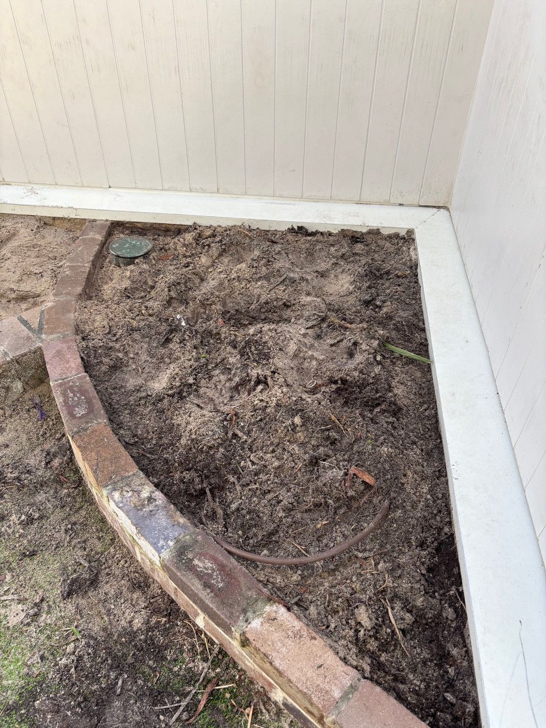 Dirt and mulch bed against a white wall and brick border, with a section of exposed brick — Rohan's Maintenance in Robina, QLD