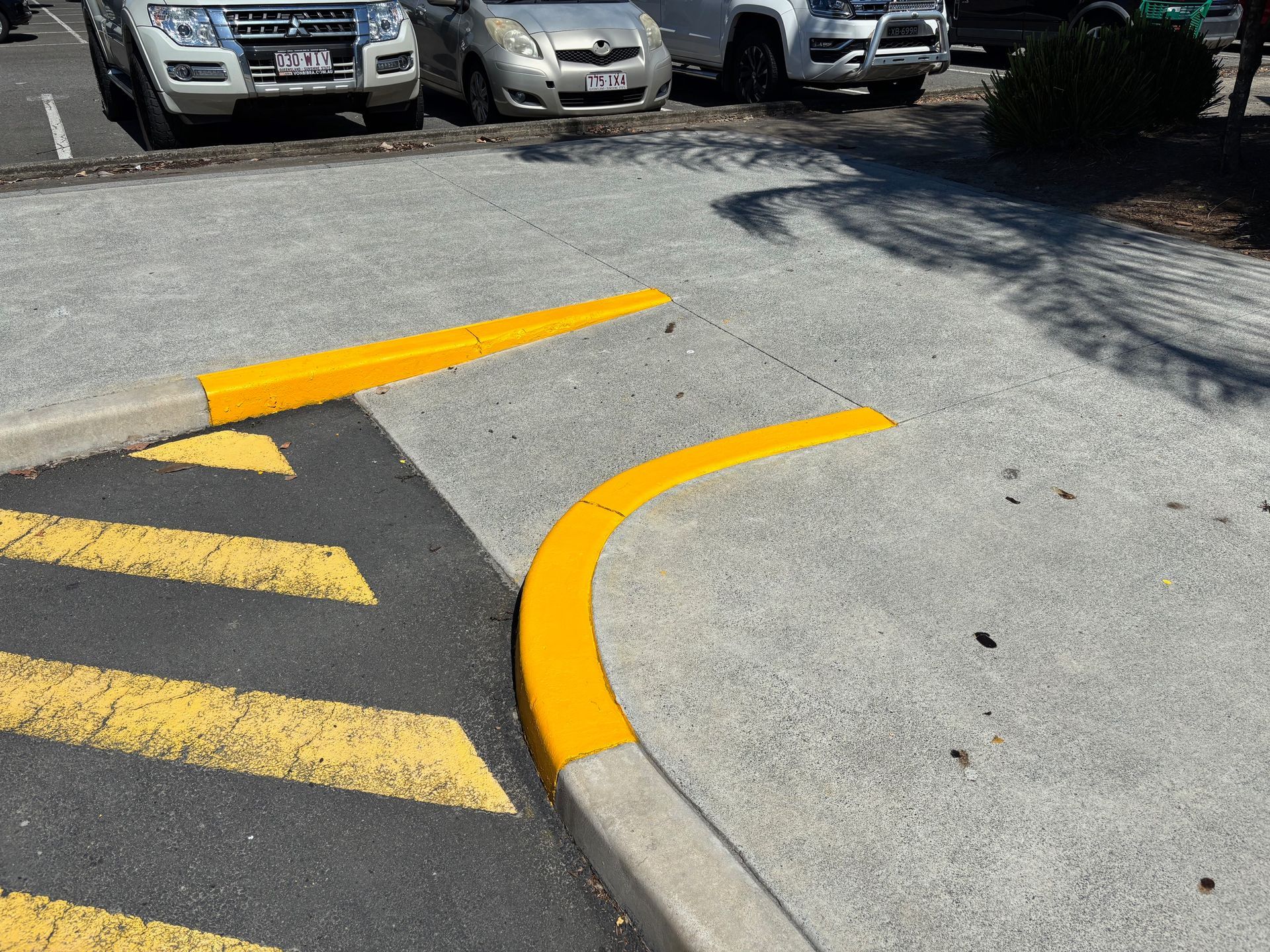Yellow curb markings at a parking area entrance, painted on concrete. Cars parked in background — Rohan's Maintenance in Robina, QLD