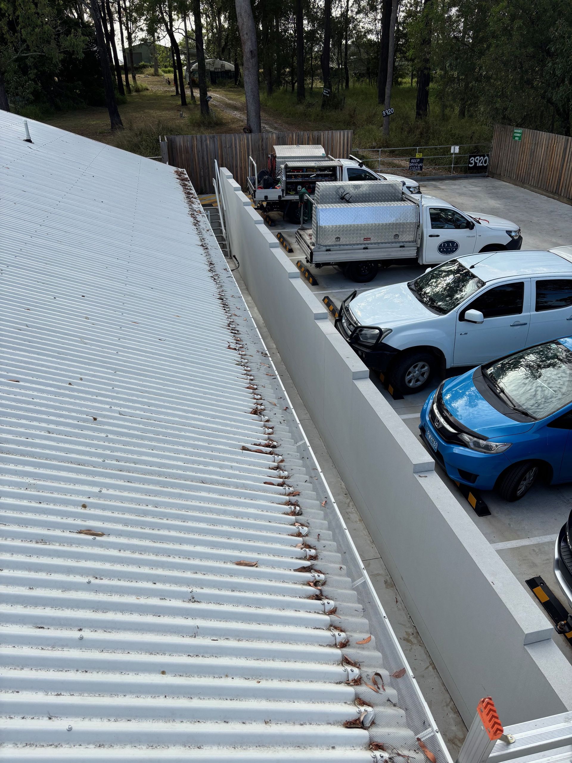 Corrugated metal roof with debris. Cars parked next to a concrete wall in a parking lot. Trees in the background— Rohan's Maintenance in Robina, QLD