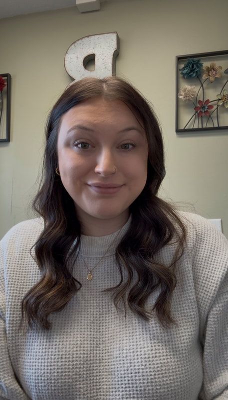 Woman with wavy brown hair smiles at the camera, wearing a light-colored sweater.