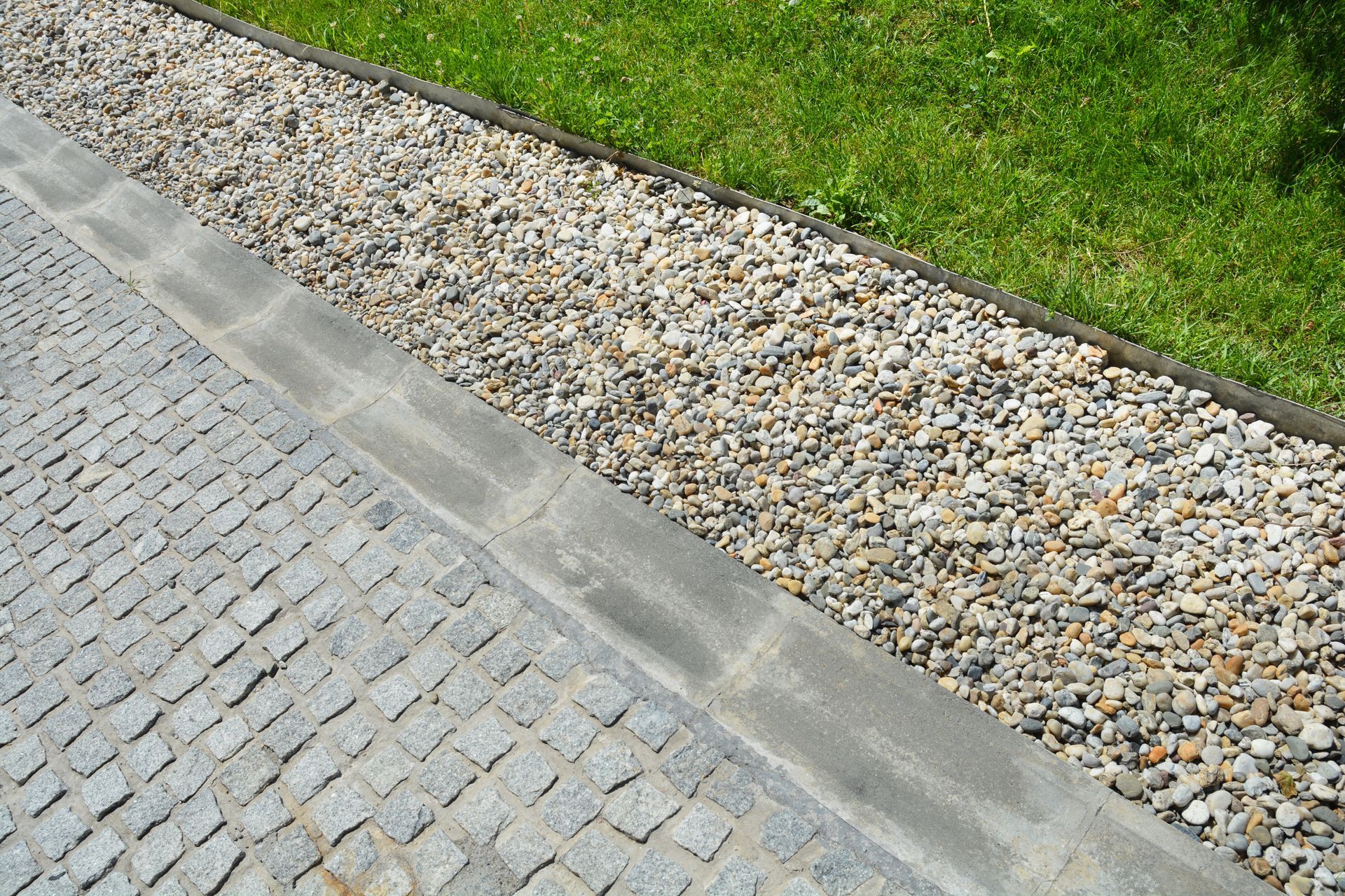 Gravel drainage bed beside stone pavement and grass lawn.