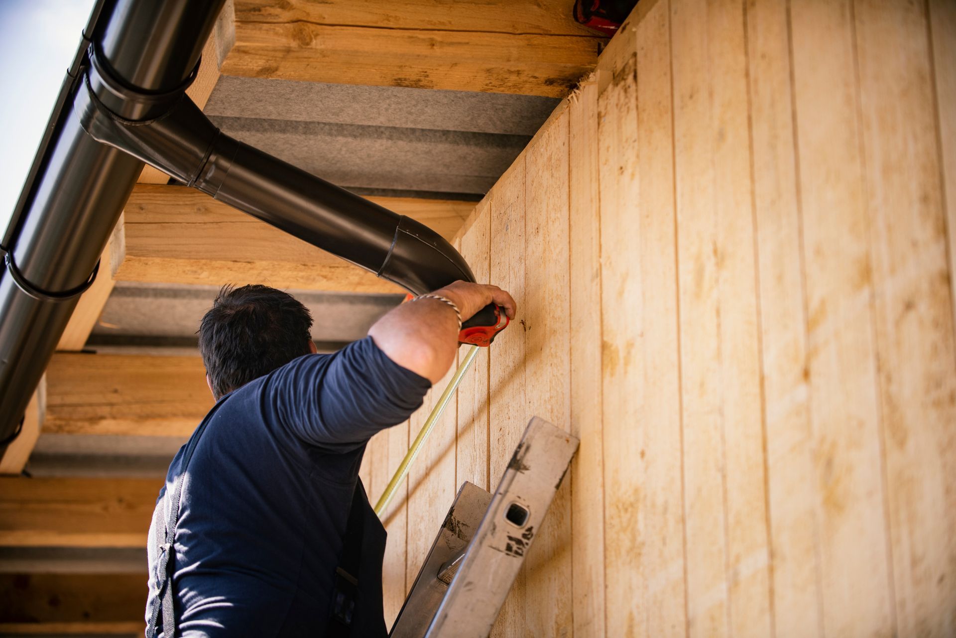 Man on ladder measuring wooden wall with tape measure under black pipe.