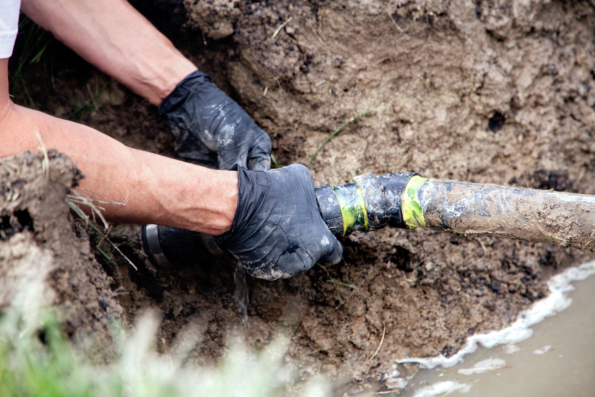 Utility worker repairing an underground drainage pipe beside a concrete slab and exposed soil.