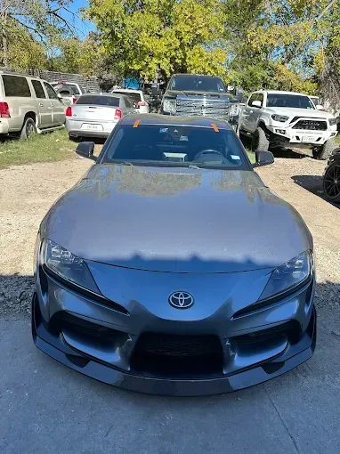 A gray Toyota Supra sports car parked outside on a sunny day with other vehicles visible.