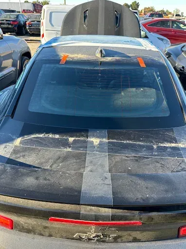 A damaged gray Chevrolet Camaro in a salvage yard, rear view with racing stripes and open hood.