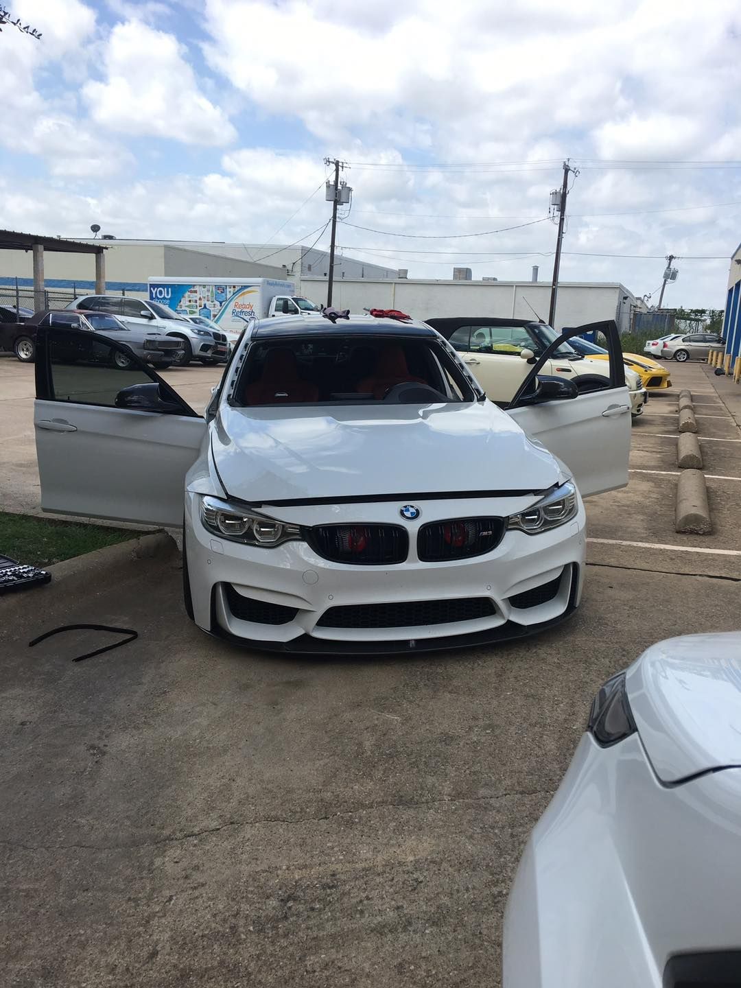 White BMW M3 with doors open, parked in front of a shop on a cloudy day.