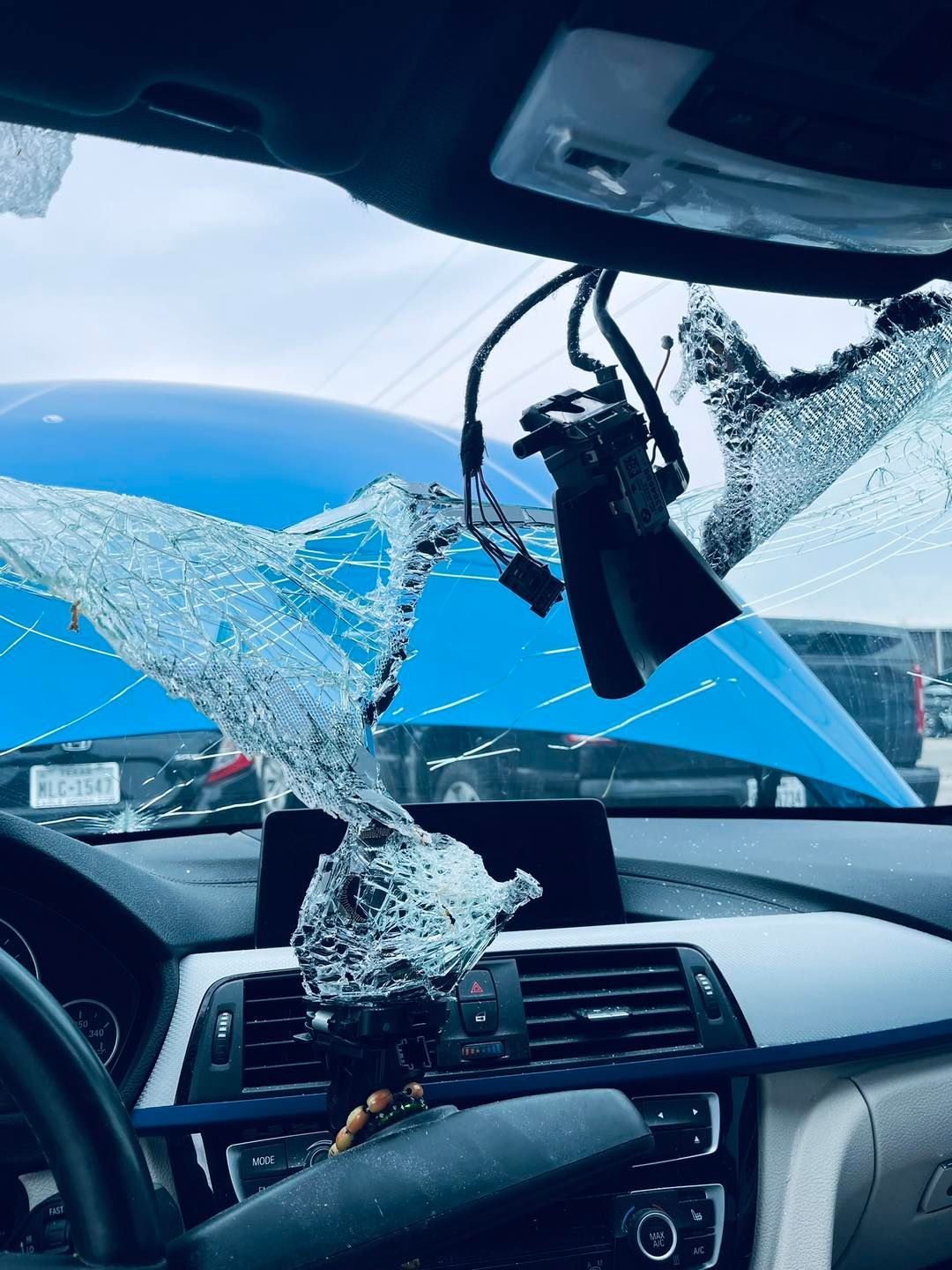 Interior view of a vehicle with a shattered windshield; dashcam hanging. Light blue car in the background.