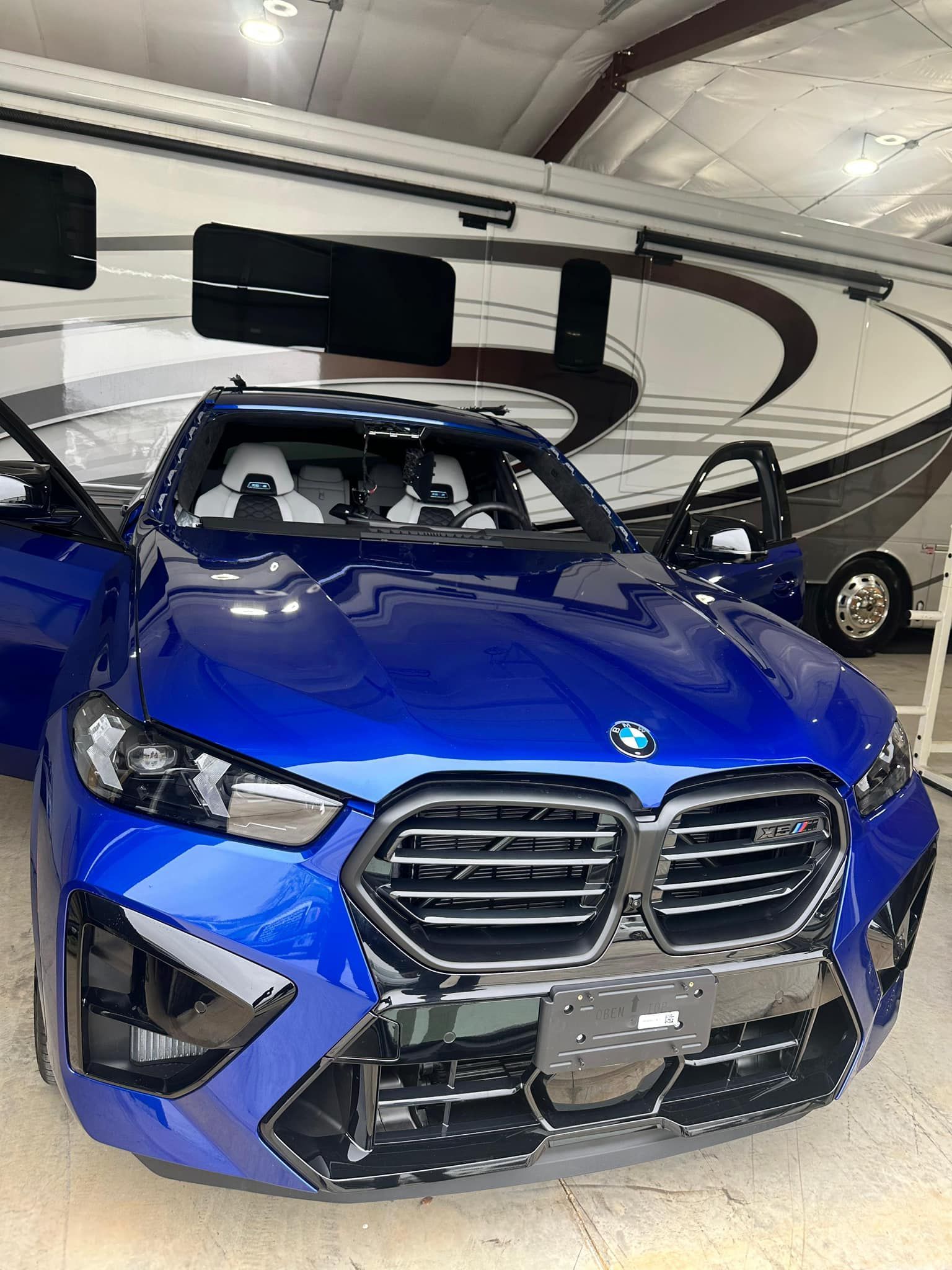 Blue BMW X6 M SUV with doors open, in front of a large RV, in a hangar.
