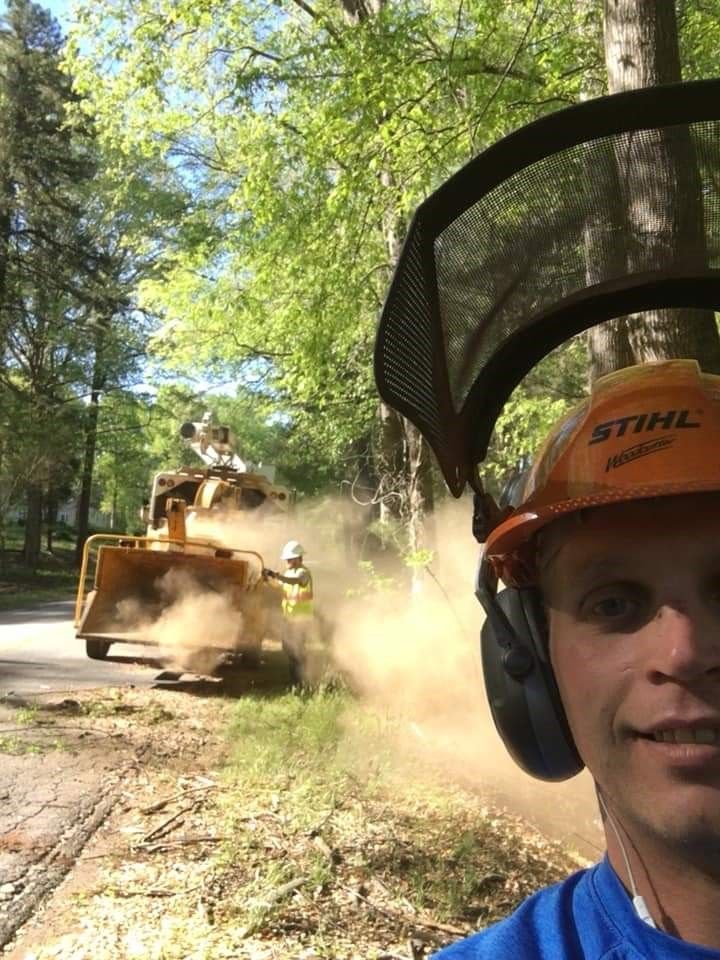 A man wearing a hard hat and headphones is taking a selfie in front of a tree chipper.