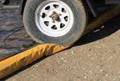 A trailer tire rolls over a yellow water-filled barrier on a dirt road