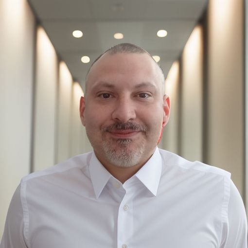 A man with a beard wearing a white shirt is smiling in a hallway