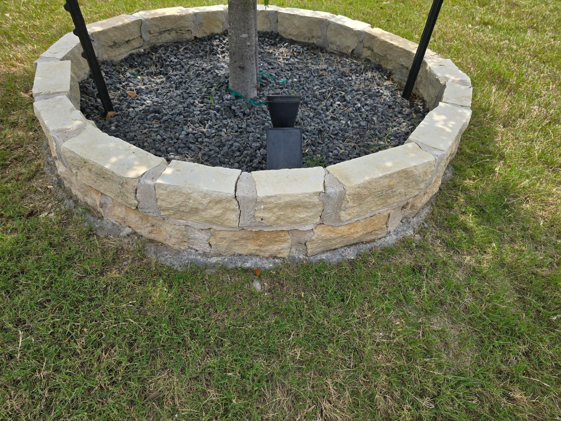 Stone-edged tree ring with dark gravel around a tree, supported by black stakes, in a grassy yard.