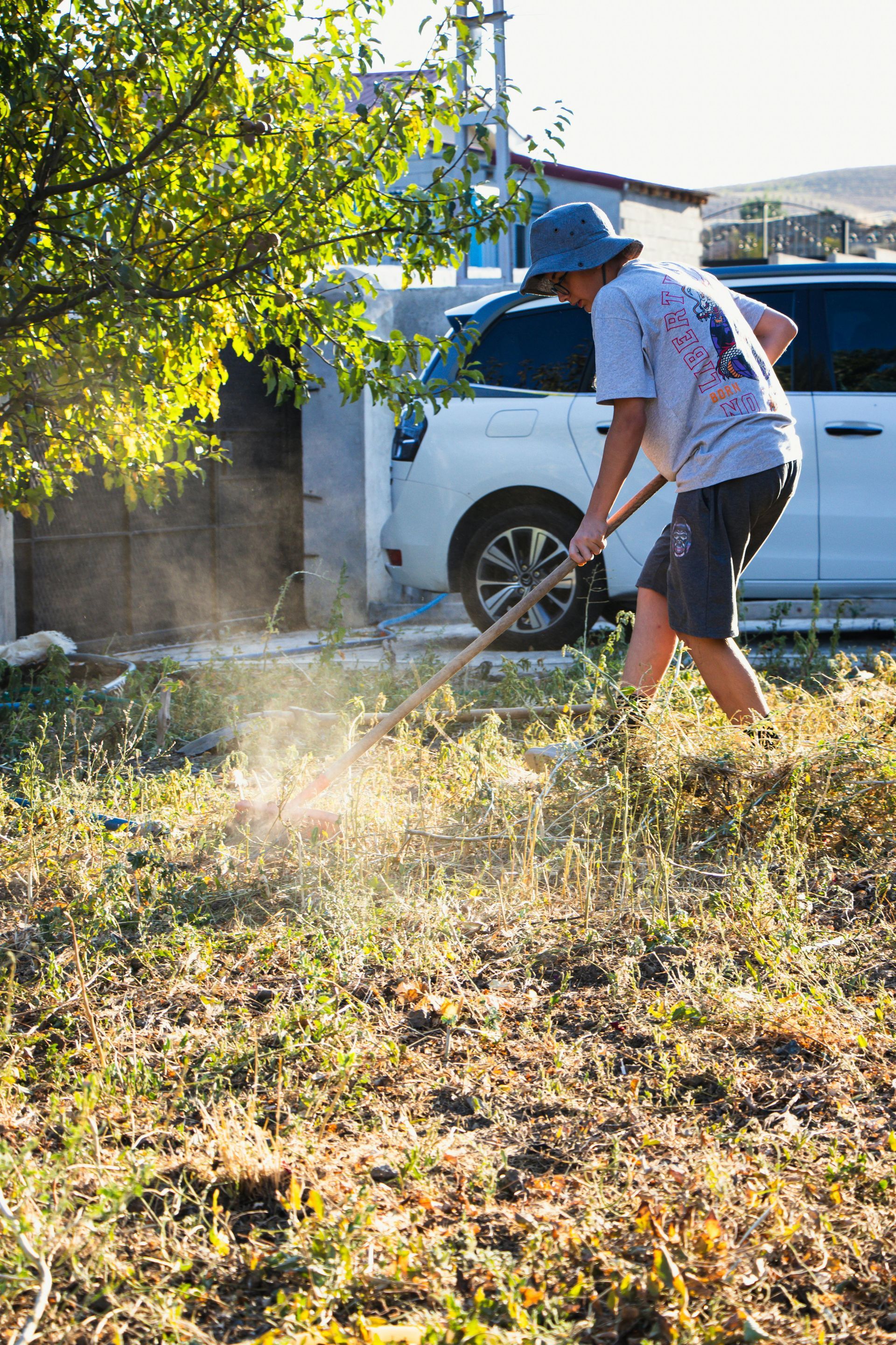 Person raking dry grass in yard near a white car and a building under a sunny sky.