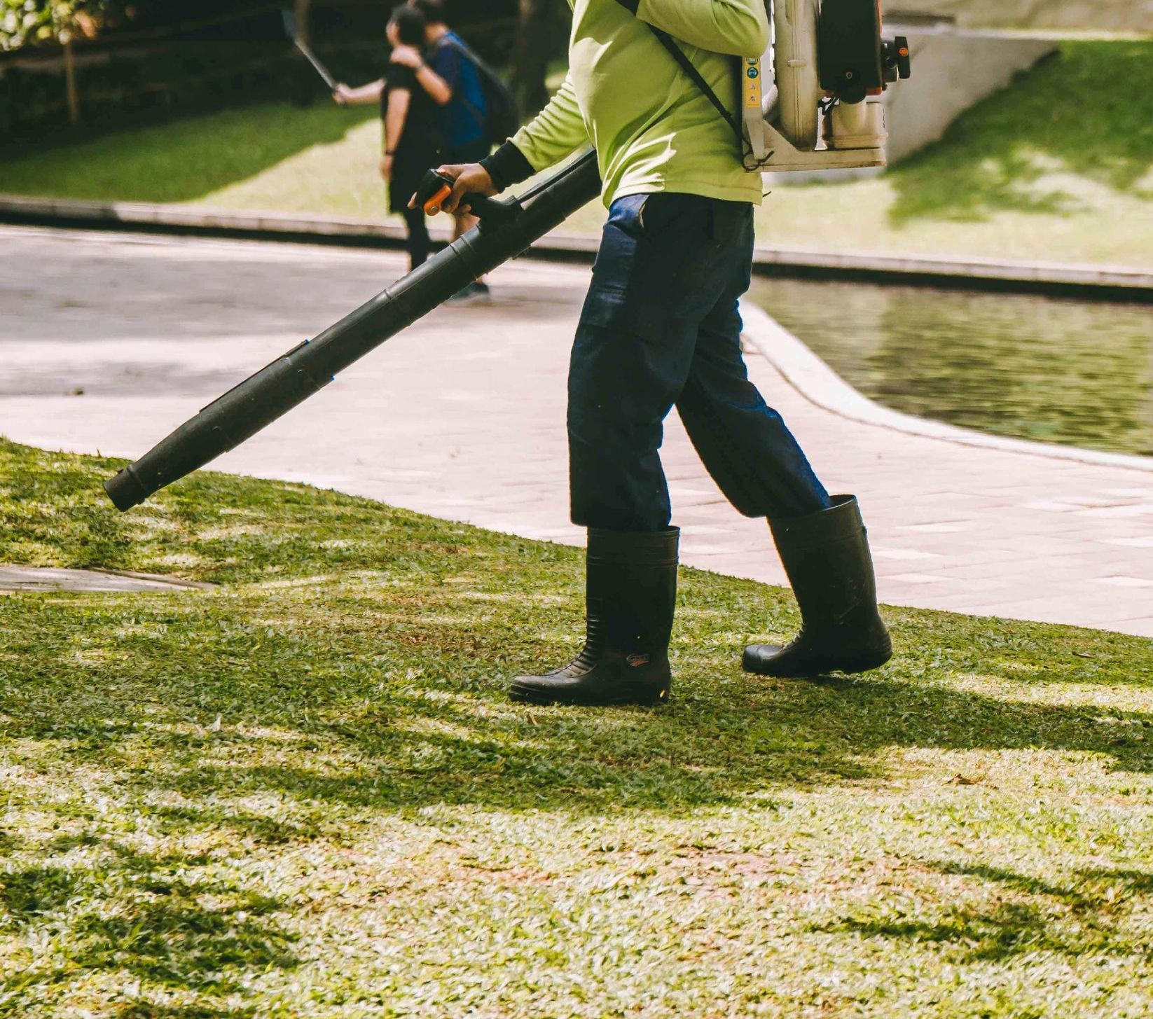 Person using a leaf blower on a grassy area near a water feature.