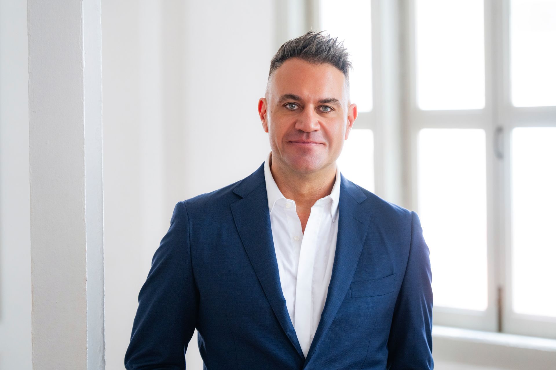 Man in blue blazer and white shirt, smiling, standing in front of a window.