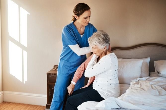 Nurse assisting an older person to sit up on a bed. The person has their hand on their neck.