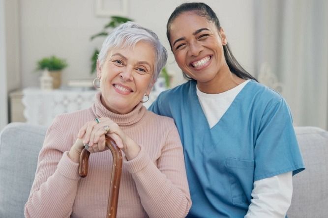 Woman with gray hair and cane smiles with caregiver in blue scrubs, indoors.