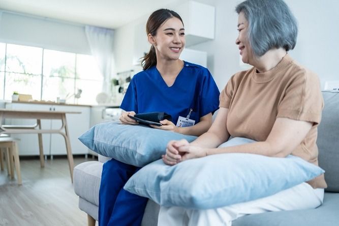 Caregiver in blue scrubs smiles at senior woman on a couch. Living room setting.